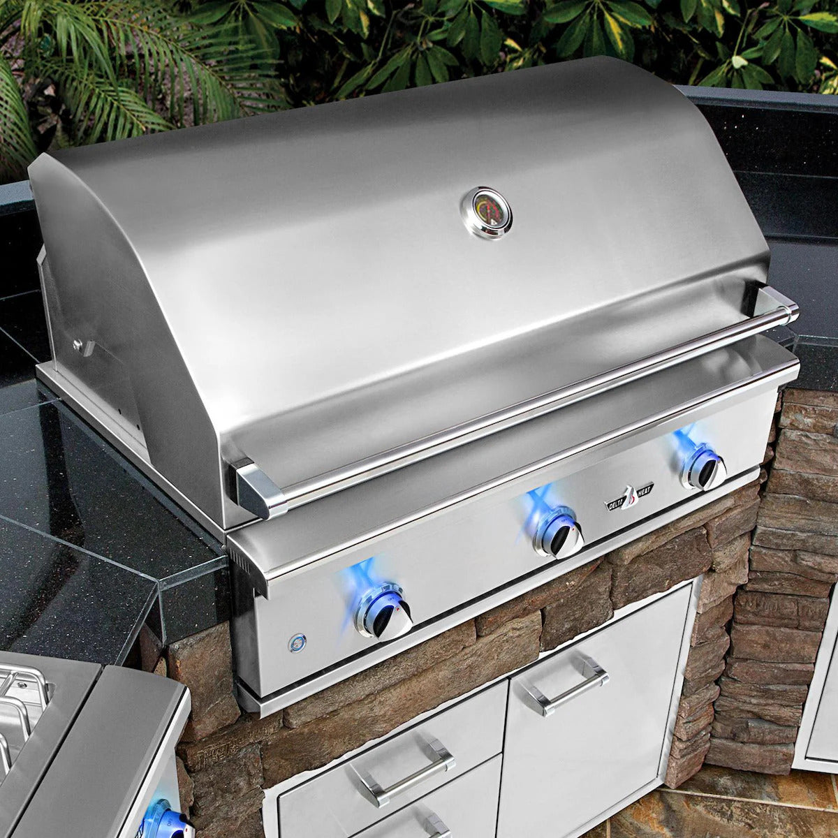 Stainless steel grill on a stone outdoor kitchen counter with greenery in the background