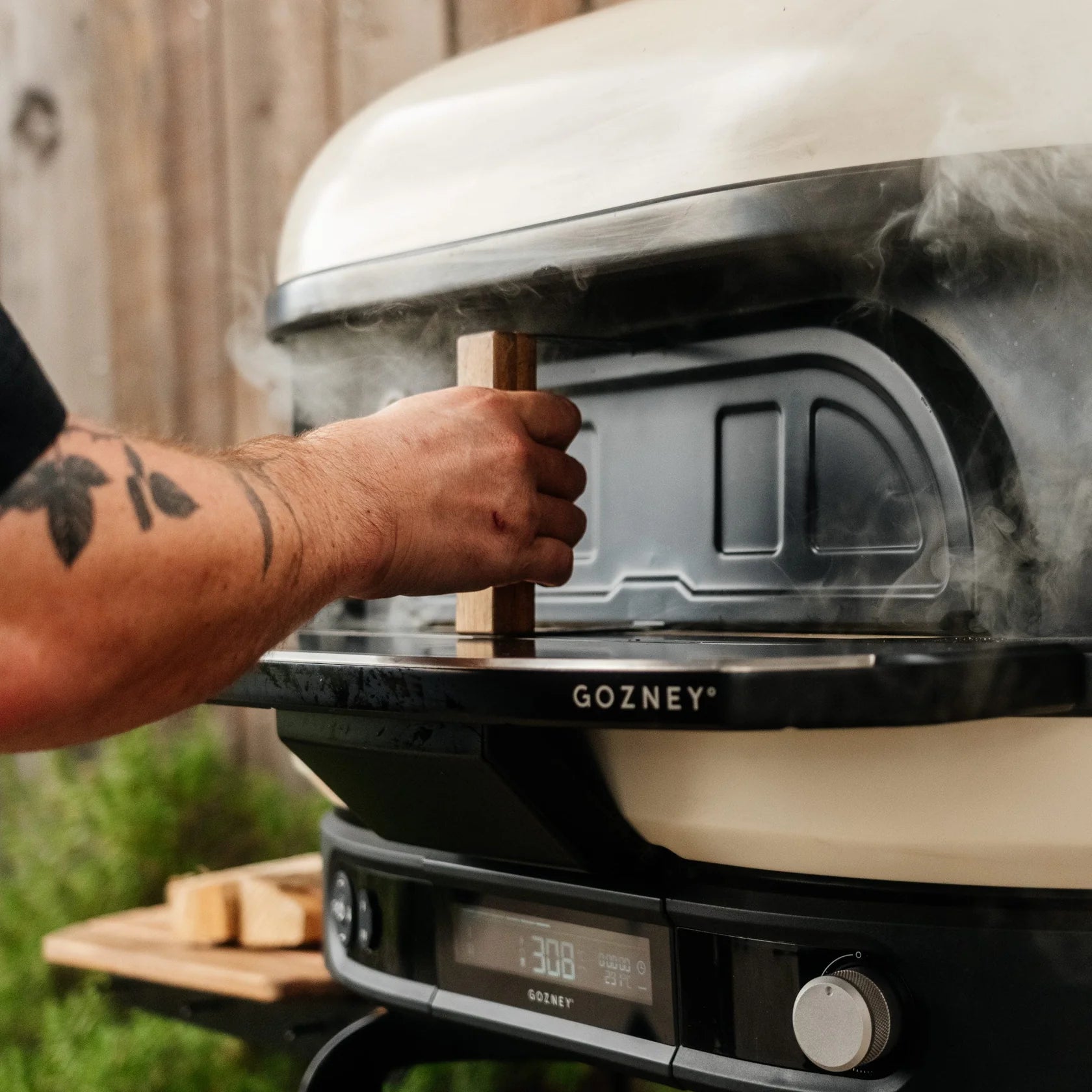 Person using a GOZNEY Gen 2 Door on a ceramic wood-fired oven with smoke coming out, outdoor setting.