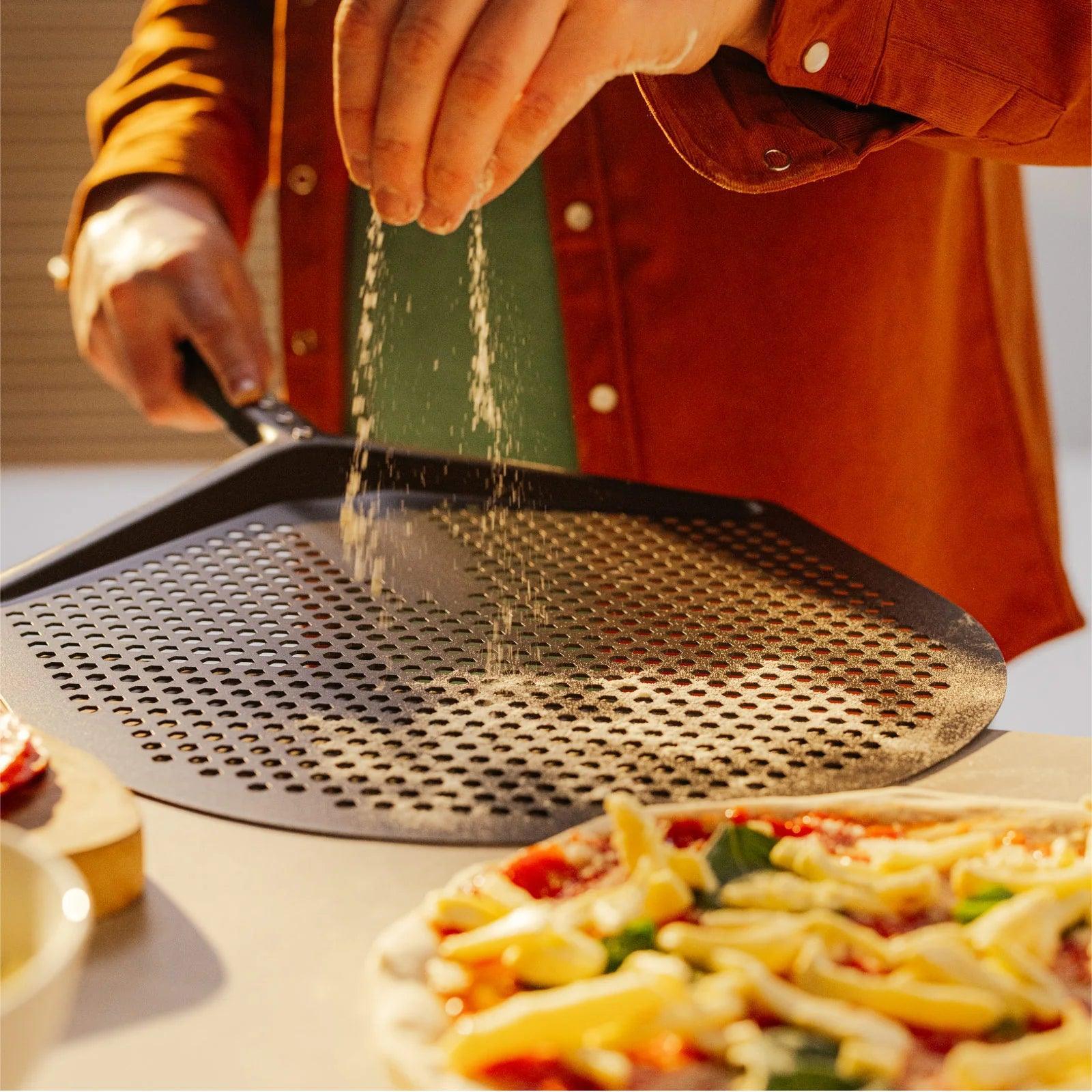 Person seasoning a pizza with grated cheese using a perforated metal pan.