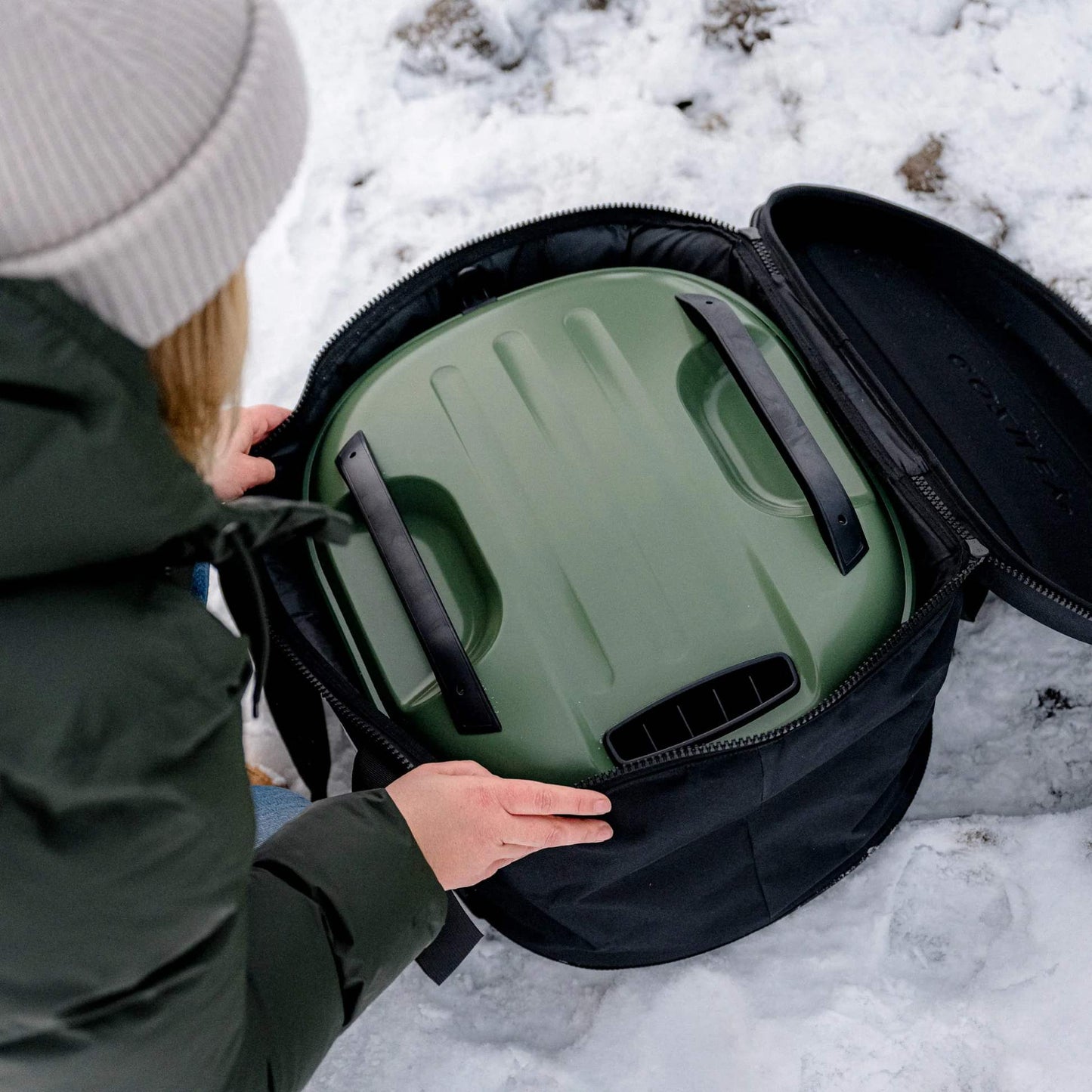 Person opening a Black Tread Venture bag with 'GOZNEY' with a green pizza oven inside on a snowy ground.